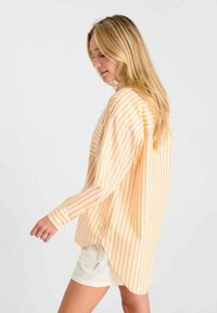 Femme aux longs cheveux blonds portant une chemise à rayures jaunes et blanches à manches longues et un short blanc, marchant devant un fond uni.