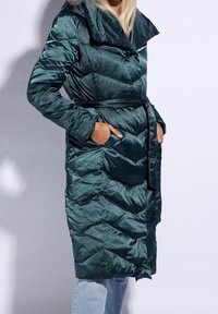 Abrigo trench acolchado verde con cinturón en la cintura, con un gran cuello y costuras en forma de chevron. Fabricado en material brillante y suave.