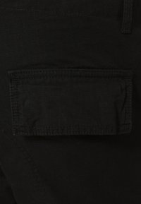 Gros plan d'une poche en tissu noir avec rabat et coutures visibles sur un matériau sombre texturé.