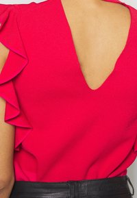 Fuchsiový top s nařasenými rukávy a hlubokým výstřihem na zádech, vyrobený z texturované látky. Na spodní straně je viditelný černý pás.