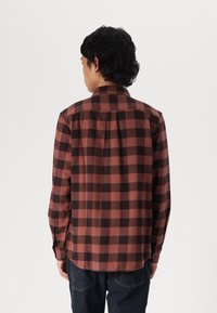 Camisa de cuadros marrones y negros con botones, de manga larga, bolsillo en el pecho, dobladillo recto y detalle de pliegue en la parte posterior. Fabricada en tejido de franela.
