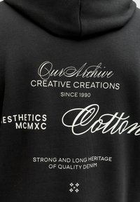 Mustast puuvillasest kangast kapuutsiga dressipluus, millel on valge tekst erinevates fontides: "Our Archive", "Creative Creations" ja "Strong Heritage of Quality Denim".