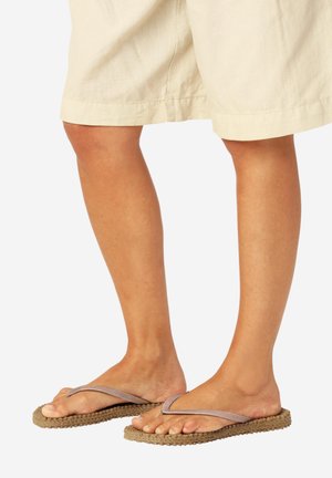 Ben iført beige shorts og brune flettede flip-flops med beige stropper, stående på en hvid baggrund.