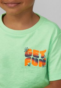 Lichtgroene katoenen t-shirt met een ribbelboord. De voorkant toont "GET FUN" in vet oranje naar donkerblauwe verloop met zon- en surfplankillustraties.