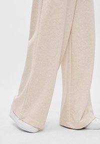 Μπεζ παντελόνι σε φαρδιά γραμμή από μαλακό ύφασμα, με άνετη εφαρμογή και λεπτή υφή. Συνδυασμένο με λευκά αθλητικά παπούτσια, σε ουδέτερη φόντο.