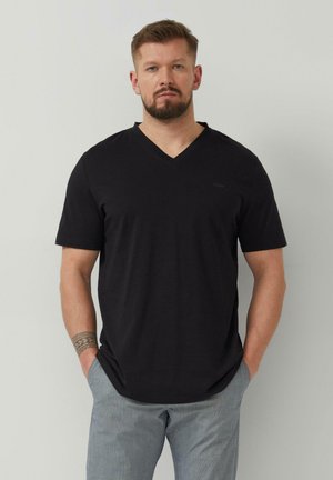 Mann mit Bart, der ein schwarzes V-Ausschnitt-T-Shirt und graue Hose trägt, steht mit den Händen in den Taschen vor einem einfarbigen Hintergrund.