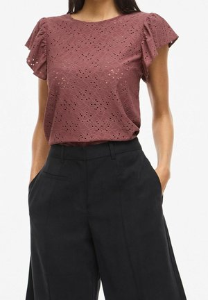 Femme portant un chemisier en dentelle ajourée mauve à manches courtes et un pantalon noir taille haute à jambes larges, les mains dans les poches.