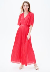 Vestido maxi rojo con pliegues, escote en V, mangas cortas y cintura ajustada con un cinturón metálico. La falda cae hasta los tobillos.