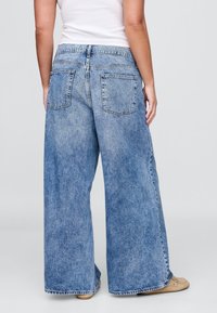 Široke modre kavbojke iz jeansa z izpiranjem, dvema zadnjima žepoma in raztrganim robom. Tkanina izgleda mehka in priložnostna.