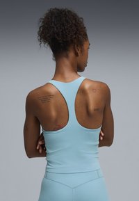 Γυναίκα με σγουρά μαλλιά, φορώντας ανοιχτό μπλε τοπ τύπου racerback και κολάν, που δείχνει πολλά μικρά τατουάζ στην άνω πλάτη και τα χέρια της.