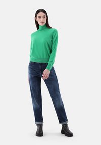 Grüner Rollkragenpullover mit langen Ärmeln, kombiniert mit blauen Jeans und schwarzen Knöchelschuhen. Die glatte Textur und der figurbetonte Schnitt betonen die Silhouette.