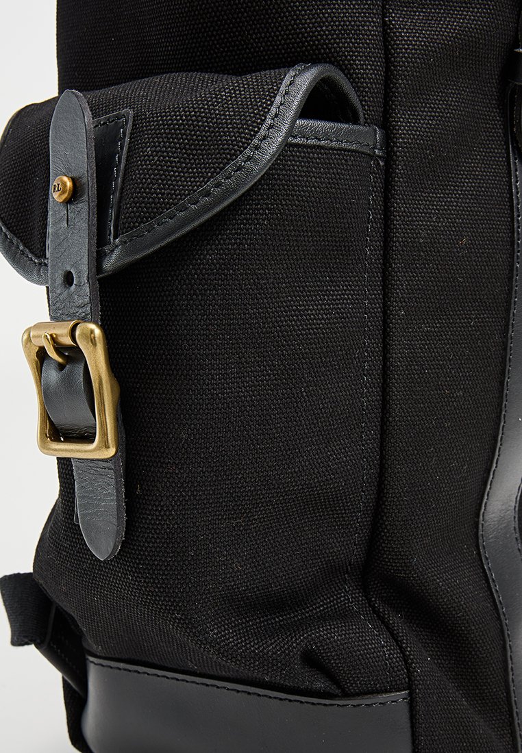 Schwarzer Canvas-Rucksack mit Lederbesatz, Messing-Schnallenverzierung und einer seitlichen Tasche mit Klappe. Strukturierte Oberfläche.