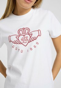 Camiseta blanca de algodón con un diseño gráfico rojo que presenta manos sosteniendo un corazón y el texto "LEE ESTD 1889" debajo del corazón.