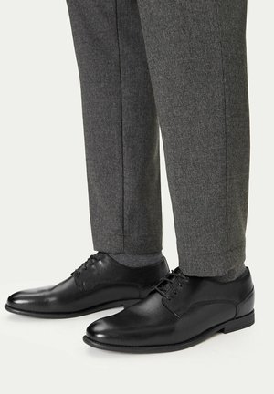 Schwarze, geschnürte Lederschuhe, kombiniert mit dunkelgrauen Hosen und passenden Socken vor hellem Hintergrund.