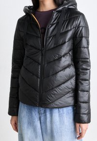 Czarna puchowa kurtka z kapturem, ozdobiona pikowaniem w jodełkę, z zamkiem błyskawicznym z przodu i kontrastowym kolorem wnętrza. Odłączane troczki kaptura.