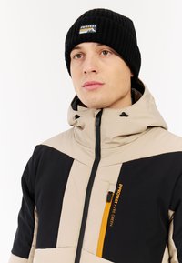Beige en zwarte jas met een ritsborstzak en een oranje accent. Draag met een zwarte geribbelde beanie met een logo patch.