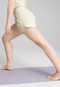 Beige sporta šorti ar izliekumu apakšmalā, pagatavoti no viegla materiāla, ar zelta logo un valkāti kopā ar atbilstošu topu uz jogas paklāja.