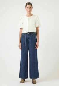 Bílá halenka s nafouklými rukávy na knoflíky v kombinaci s tmavě modrými džínami širokých kalhot. Modelka má na sobě hnědé podpatky. Minimalistický design.