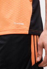 Κοντινό πλάνο ενός ατόμου που φοράει πορτοκαλί και μαύρη αθλητική στολή με το κείμενο «Climacool» στο μπλουζάκι και ριγέ σορτς, με το χέρι ορατό στο πλάι.