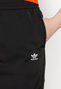 Mano appoggiata all'interno della tasca di pantaloni neri Adidas con logo bianco, maglietta arancione parzialmente visibile sopra la vita.