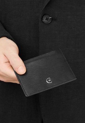 Mano sosteniendo un tarjetero de cuero negro texturizado con logo plateado sobre un fondo de tela de blazer negra con botones.