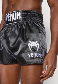 Calções de boxe em cetim preto com cintura franzida. Apresentam logótipo bordado a branco e detalhes gráficos. Fendas laterais para facilitar os movimentos.