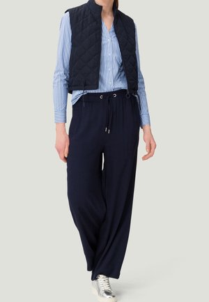 Mujer con camisa de rayas azules abotonada, chaleco acolchado azul marino, pantalones anchos azul marino con cordón y zapatillas plateadas, de pie sobre un fondo liso.