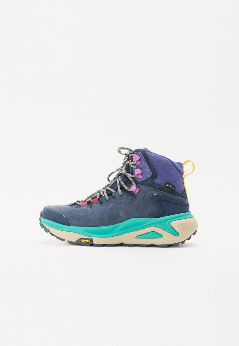 Mid-top hiking shoe in dark blue and purple, featuring a cushioned turquoise sole, pink and gray laces, and a textured upper. Vibram branding visible.