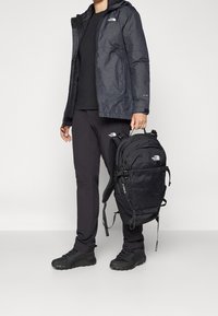 Person som bär en svart utomhusjacka, svarta byxor och skor, och håller en svart North Face ryggsäck i sitt topphandtag.