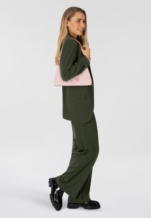 Sac à main rose matelassé avec des éléments en argent, comportant des accents en forme de cœur. Le modèle porte un costume vert olive et des chaussures à plateforme noires.