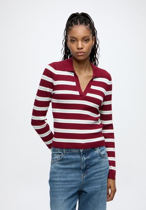 Woman with braided hair wears a red, white, and blue striped long-sleeve shirt with a collar and blue jeans, standing against a plain background.