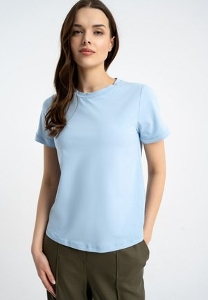 Kobieta z długimi brązowymi włosami, ubrana w jasnoniebieską koszulkę z krótkim rękawem i oliwkowo-zielone spodnie, stojąca na białym tle.