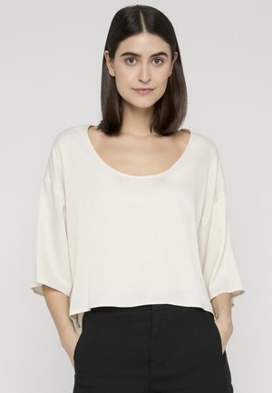 Frau mit glattem dunklem Haar, die ein lockeres weißes Oberteil mit rundem Ausschnitt und schwarze Hosen trägt, mit den Händen in den Taschen vor einem einfarbigen Hintergrund stehend.