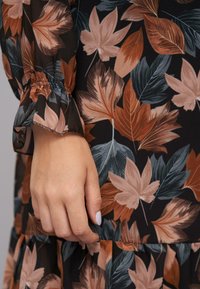 Bloemenuur met bruine, beige en groene bladerenpatronen op een zwarte achtergrond. Kenmerken zijn onder andere gebloemde mouwen en geplooide stof.