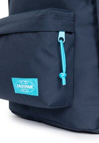 Zaino in tessuto blu navy con una tasca anteriore dotata di una zip azzurro chiaro e un'etichetta blu acqua con il logo EASTPAK. Superficie testurizzata.