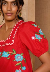 Blusa rossa con ricami floreali in blu e rosa, scollatura quadrata e maniche a sbuffo. Orecchini circolari dorati completano il look.