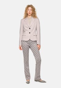 Hellgrau maßgeschneiderte Blazer mit zwei Knöpfen und Taschen, kombiniert mit gemusterten Hosen, die kreisförmige Designs in Grautönen und Beige aufweisen.