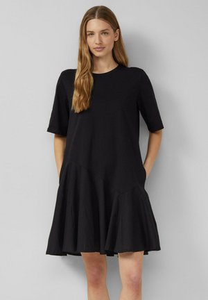 Frau mit langen Haaren, die ein locker sitzendes schwarzes Kurzarmkleid mit ausgestelltem Rock trägt und vor einem einfarbigen hellgrauen Hintergrund steht.