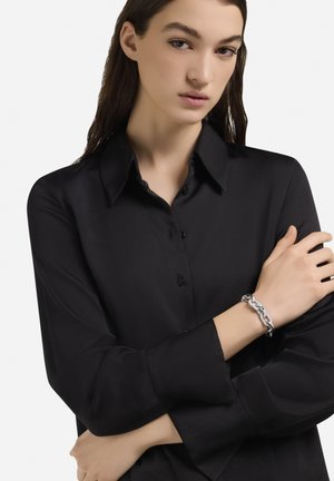 Zwart satijnen overhemd met lange mouwen en een spitse kraag, aangevuld met een zilveren armband aan de rechterpols. Gladde textuur, nauwsluitend ontwerp.