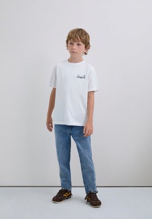 T-shirt bianca in cotone con maniche corte e scollatura rotonda, con testo ricamato blu navy. Indossata con jeans azzurri e sneakers marroni.