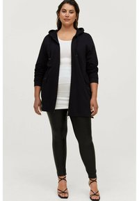 Langer schwarzer Kapuzenmantel mit Reißverschlusstaschen, kombiniert mit einem taillierten weißen Oberteil und glänzenden schwarzen Leggings. Schwarze Riemchensandalen runden den Look ab.