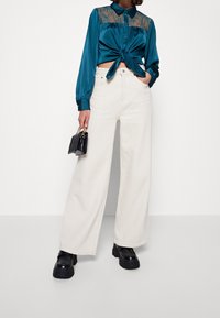 Tealfärgad satin skjorta med långa ärmar, spetsdetaljer och knut fram, kombinerad med vida krämfärgade byxor och kraftiga svarta plattformsskor.