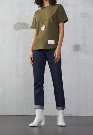 Person iført olivengrøn kortærmet skjorte, mørkeblå opsmøgede jeans, hvide ankelstøvler og en lang vedhængskæde står på gråt gulv.