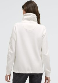 Frau in einem weißen Sweatshirt mit hohem Kragen und erhabenem Text am Kragen und Ärmel, kombiniert mit dunklen Jeans, die mit dem Rücken zur Kamera steht.