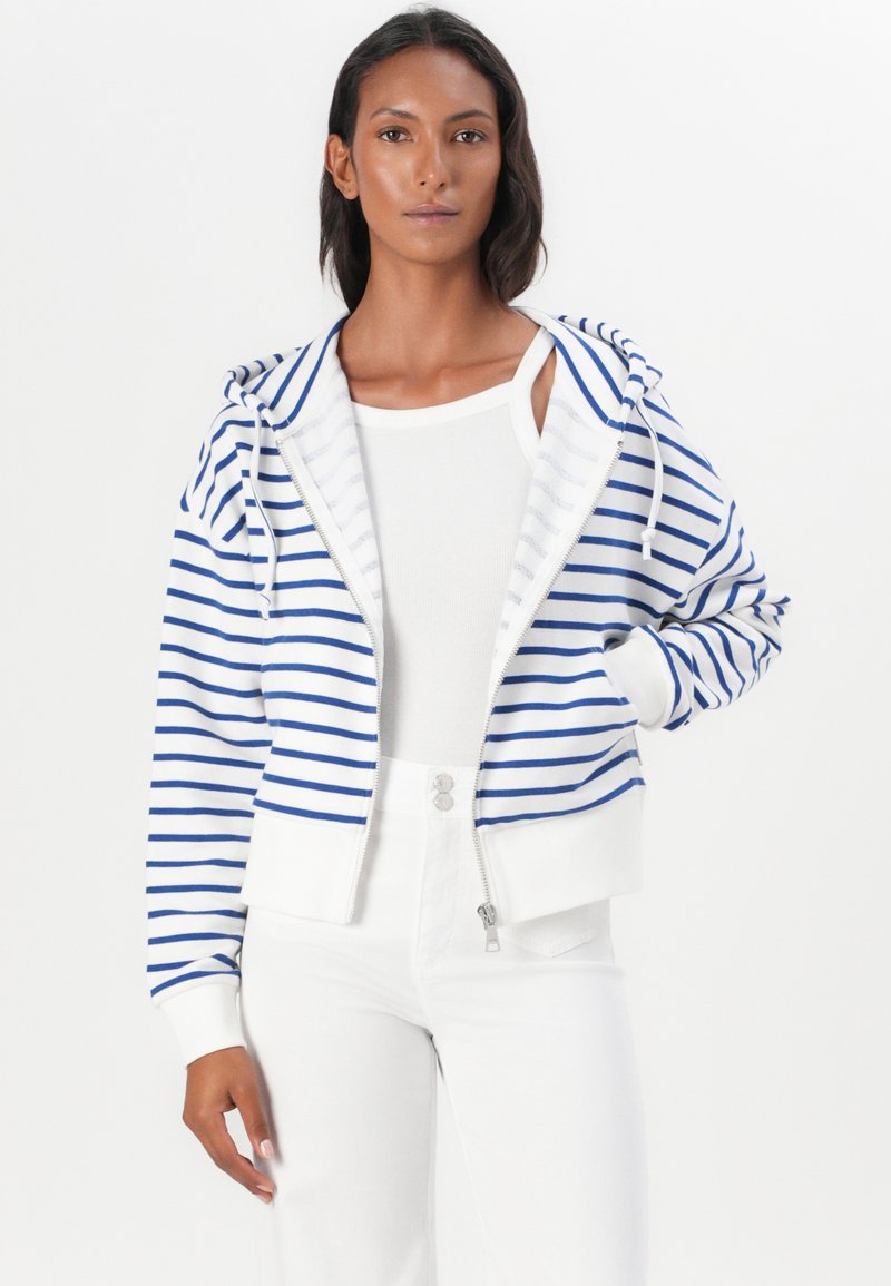 Kurtka z kapturem na zamek błyskawiczny w niebieskie i białe poziome paski, z ściągaczami przy mankietach i w pasie. W połączeniu z białym topem na ramiączkach i białymi spodniami.