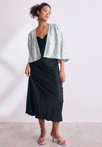 Zwarte satijnen jurk met een valkraag, gecombineerd met een sprankelend zilveren cropped jack. Strappy doorzichtige hakken maken de outfit compleet.