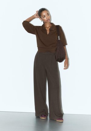 Donna che indossa un top marrone a maniche lunghe, pantaloni larghi scuri, scarpe bordeaux e porta una grande borsa a tracolla scura, in piedi con una mano sulla testa.