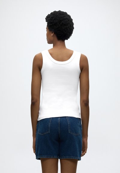 Άτομο με κοντά κατσαρά μαλλιά, που φοράει λευκό τοπ χωρίς μανίκια και σκούρες μπλε τζιν βερμούδες, με την πλάτη προς μια απλή φόντο.