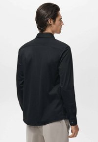 Hombre con el cabello peinado hacia atrás, viste una camisa negra de manga larga y pantalones beige, de espaldas contra un fondo claro y sencillo.