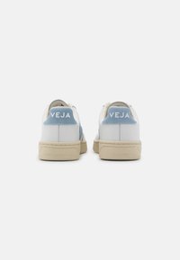 Sneakers Veja bianchi e beige con linguette in suede blu chiaro, viste da dietro su uno sfondo bianco neutro.